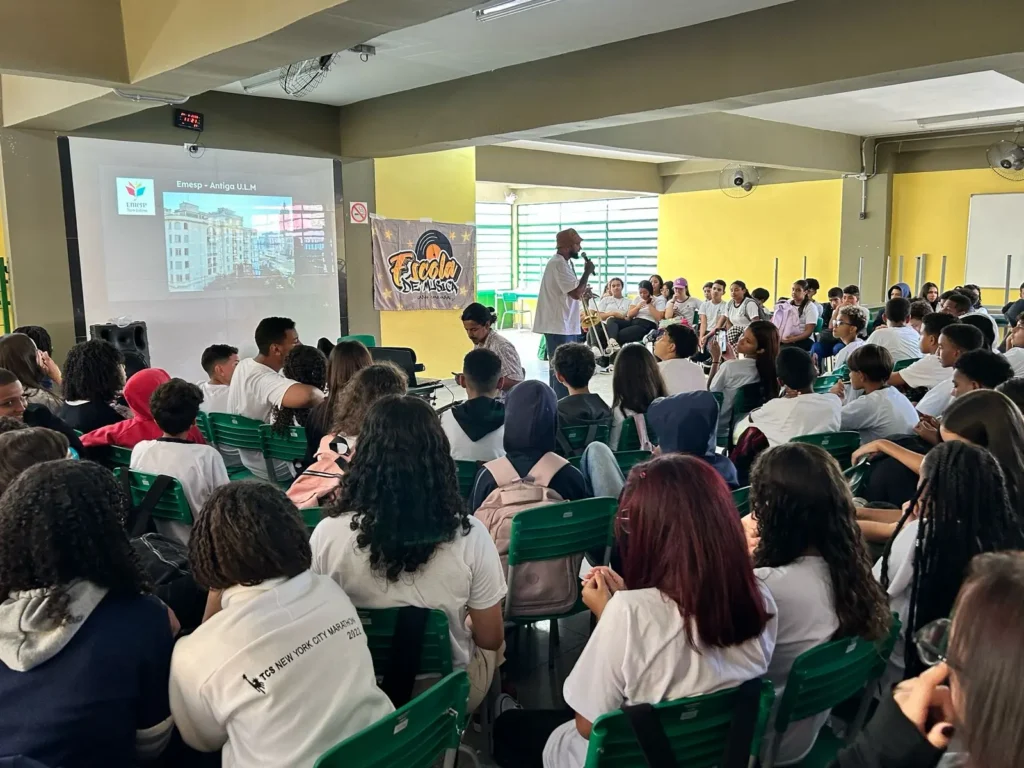 Estudantes reunidos em sala de aula durante atividade musical do projeto Capão–Congonhas, com músico segurando um trombone à frente da turma.